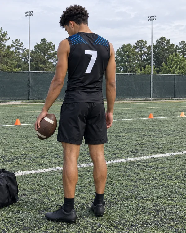 A 7v7 player seen from behind on a field, in a black sleeveless top with a white 7 and black shorts, holding a football