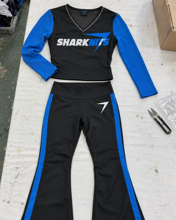 The KICKLINE uniform laid flat: a black top with blue sleeves, clear stones along the V-neck and SHARKNITS across the chest, over black flared pants with blue side panels