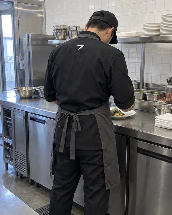 A cook seen from behind at a steel kitchen pass, in a black chef coat with a white bolt on the back and a charcoal apron
