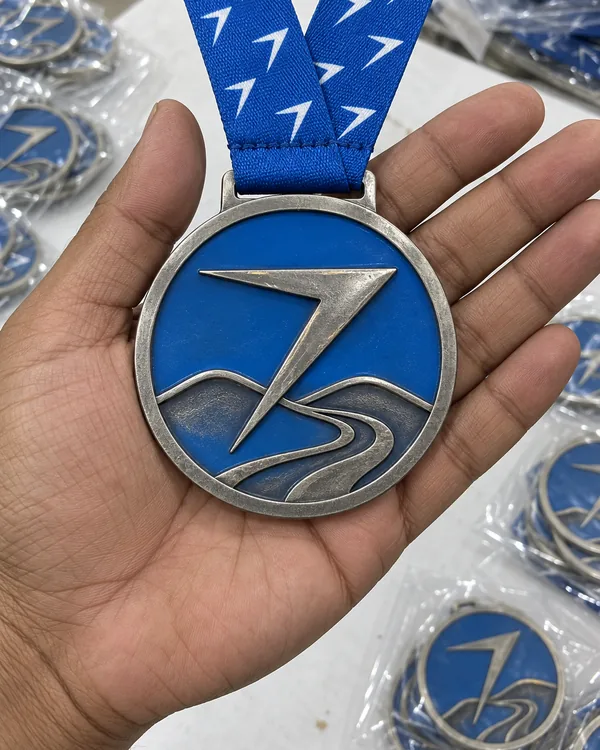 A finisher medal held flat on a palm: a raised bolt over a road winding through hills, filled in blue, on a blue ribbon printed with small white bolts