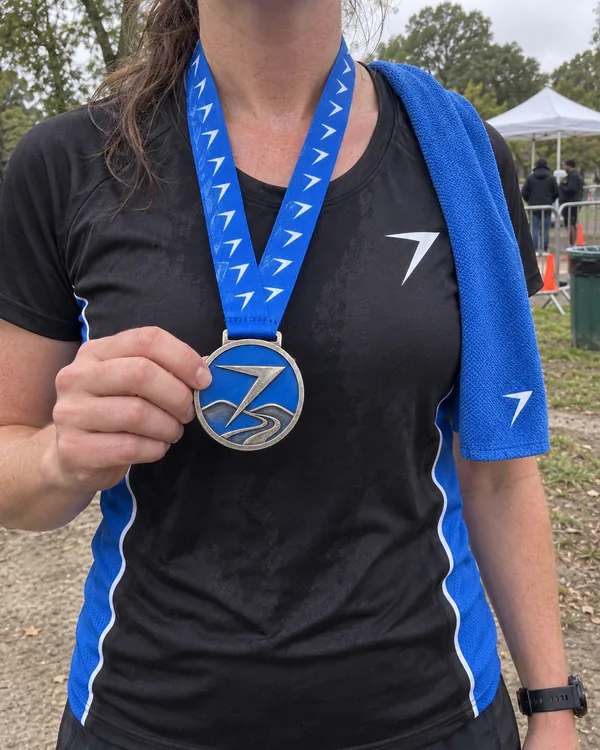 A runner after a race holds up her finisher medal, a raised bolt over a winding road in blue, wearing the black PACE race shirt with blue mesh side panels and a blue towel on one shoulder