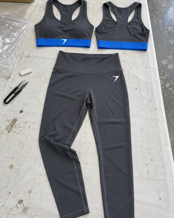 PULSE leggings and two racerback sports bras laid flat on a factory table, charcoal with a blue band