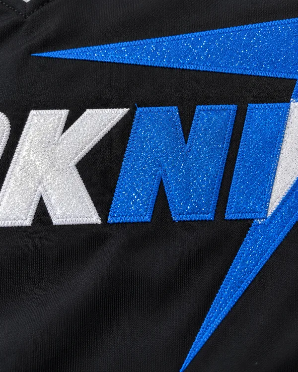 SHARKNITS letters in white and blue sparkle twill, stitched with a zigzag onto a black shell, close up