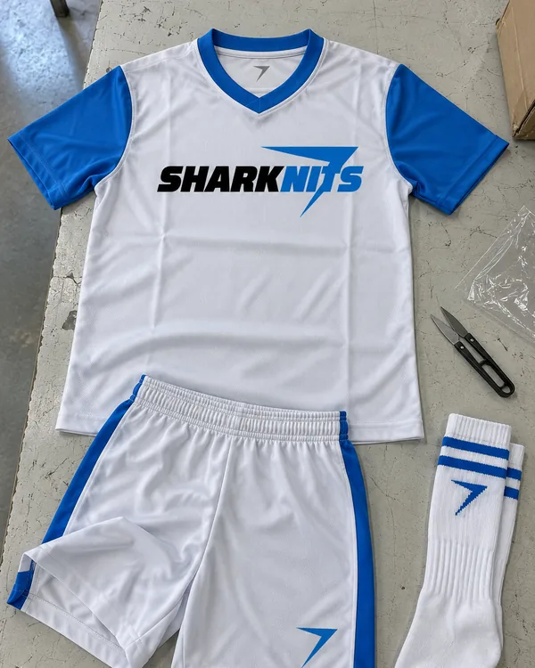 The ROOKIE youth soccer kit laid flat: a white jersey with blue sleeves and SHARKNITS across the chest, white shorts with blue side panels and a striped sock