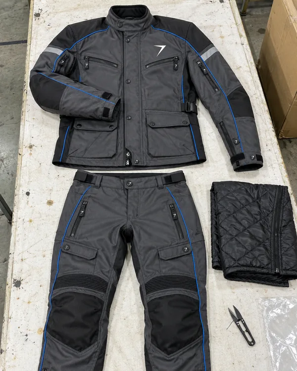 A charcoal textile riding jacket and pants with blue piping laid flat, beside a quilted liner