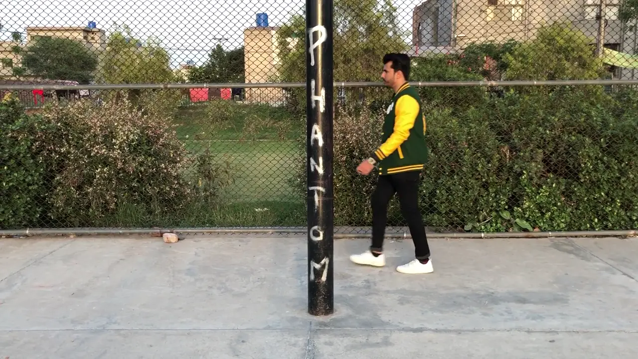A man walks past a basketball court fence in a green Fince Up varsity jacket