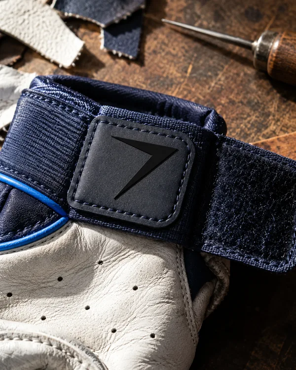 Close up of a batting glove: a navy wrist strap with a raised rubber bolt tab, blue piping and a perforated white leather palm