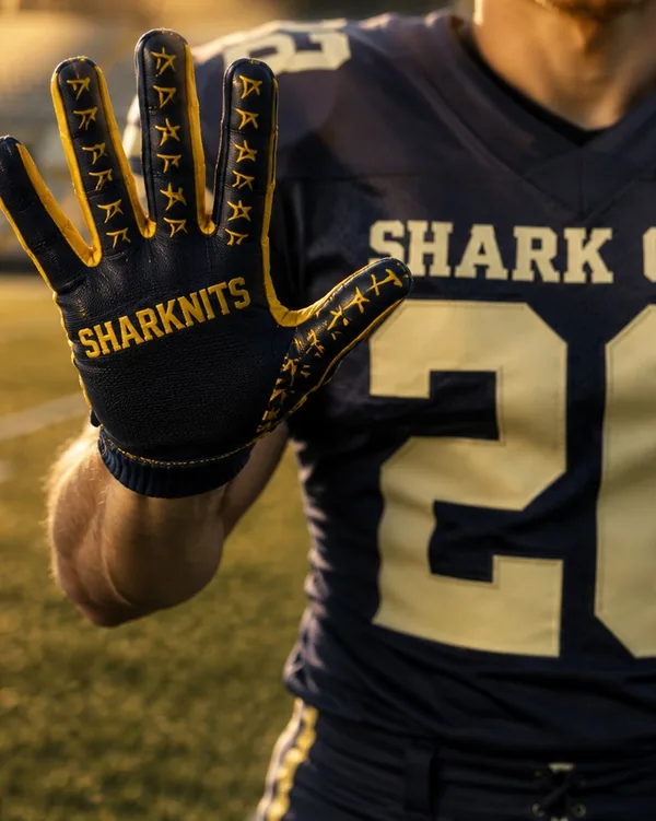 Player in a navy SHARK CITY 26 jersey holding up a black and gold football glove printed with SHARKNITS