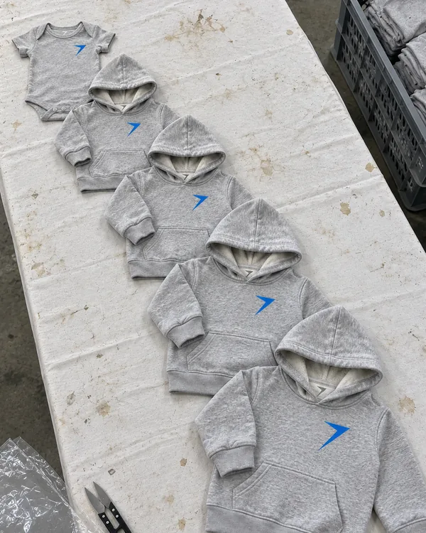 One gray hoodie design laid down a factory table in five sizes, from a baby bodysuit to a youth hoodie, each with a blue bolt