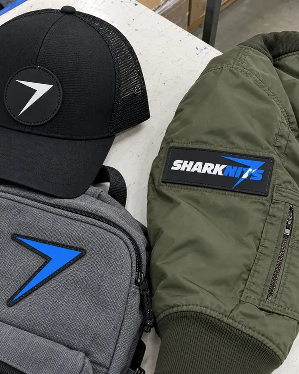 A black trucker cap, a gray backpack and an olive bomber sleeve, each with a PVC patch stitched on