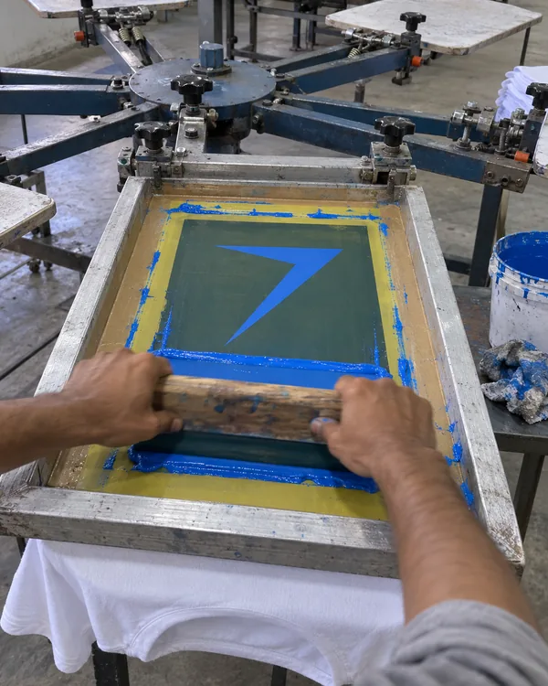 Hands pull a squeegee across a screen on a carousel press, printing a large blue bolt onto a white tee