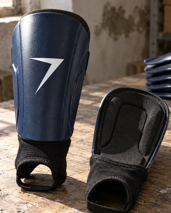 Close up of a navy shin guard with a white bolt on the shell and a black ankle sleeve, next to a guard showing its foam backing