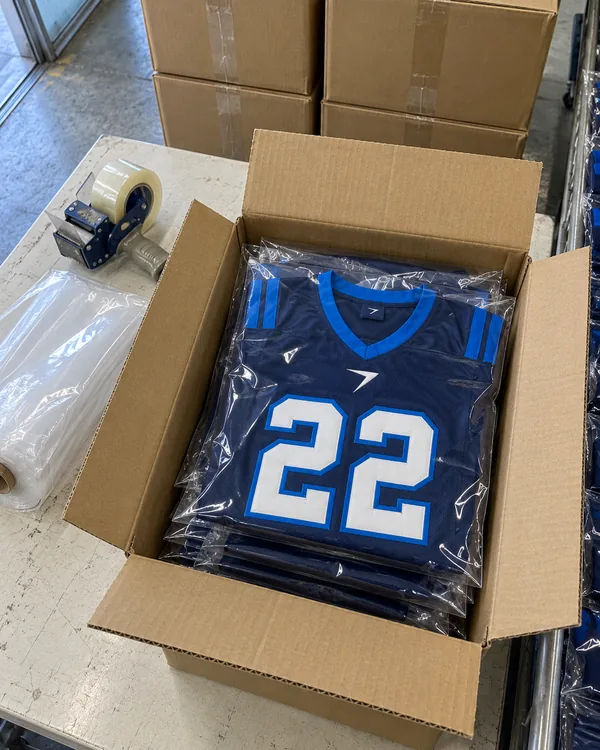Navy game jerseys with a white 22, folded in clear bags in an open carton, beside a tape gun