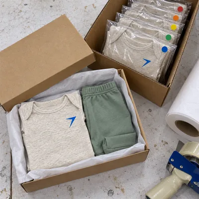 A baby set in a gift box with tissue, beside a carton of bagged bodysuits with color dot stickers
