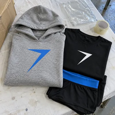 A gray hoodie with a blue bolt, a black tee with a white bolt and black shorts, folded on a packing table