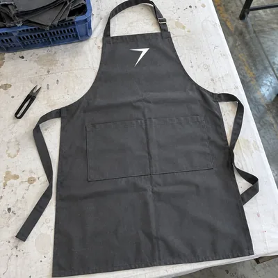 A charcoal bib apron laid flat, with a divided front pocket and a white bolt on the bib