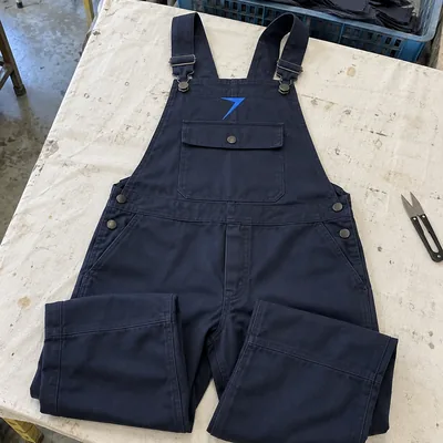 Dark navy bib overalls laid flat, with a blue bolt above the bib pocket and metal buckles on the straps