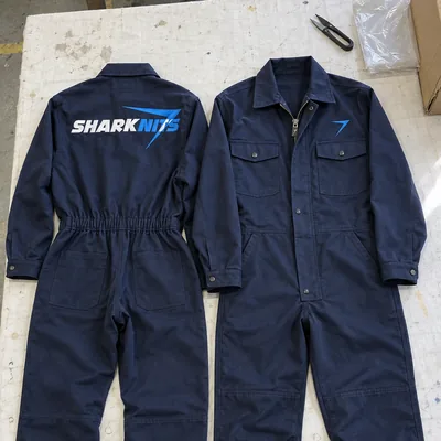 Two dark navy coveralls laid flat, one with SHARKNITS across the back and one with a blue bolt on the chest