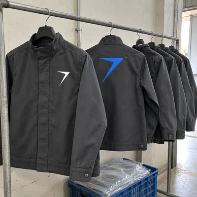 Charcoal welding jackets on a factory rail, one showing a white bolt on the chest and the next a large blue bolt on the back