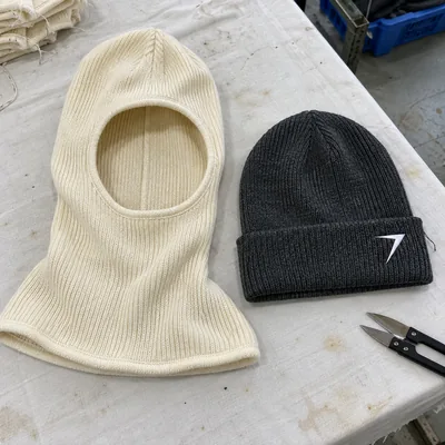 An off-white knit welding hood laid flat beside a charcoal knit hard hat liner with a white bolt on the cuff