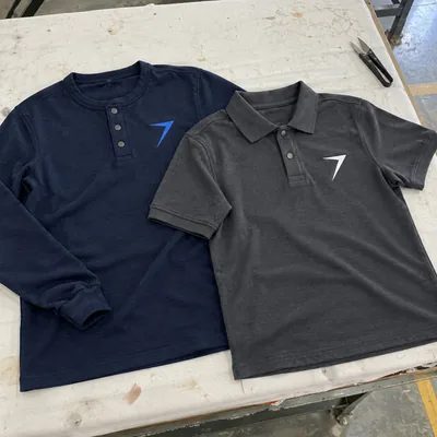 A dark navy henley with a blue bolt and a charcoal polo with a white bolt, laid side by side