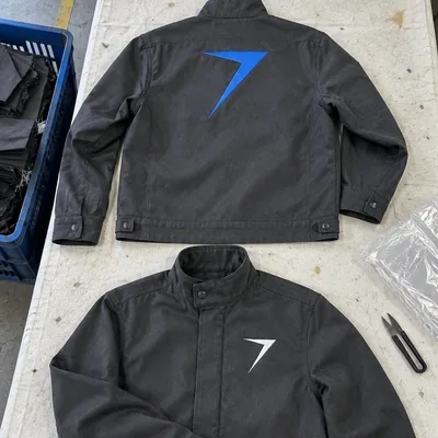 Two charcoal welding jackets laid flat, one with a large blue bolt on the back and one with a white bolt on the chest