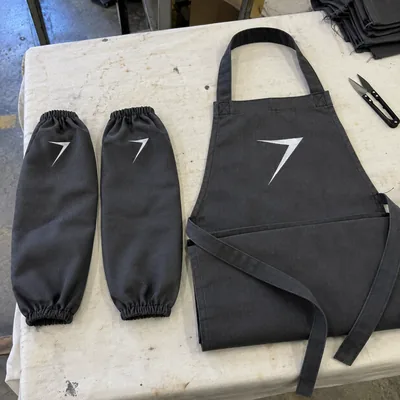 A pair of charcoal welding sleeves with elastic ends beside a charcoal bib, each with a white bolt