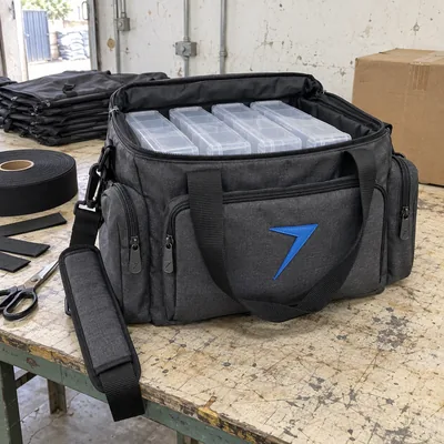 Dark gray soft tackle bag with trays inside and a blue rubber bolt patch on the front pocket