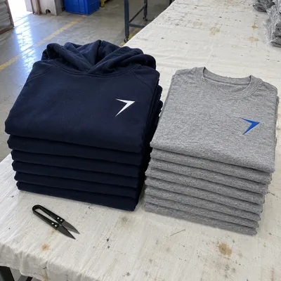 Stacks of navy hoodies and heather gray tees, each with a bolt on the chest, beside a pair of thread snips